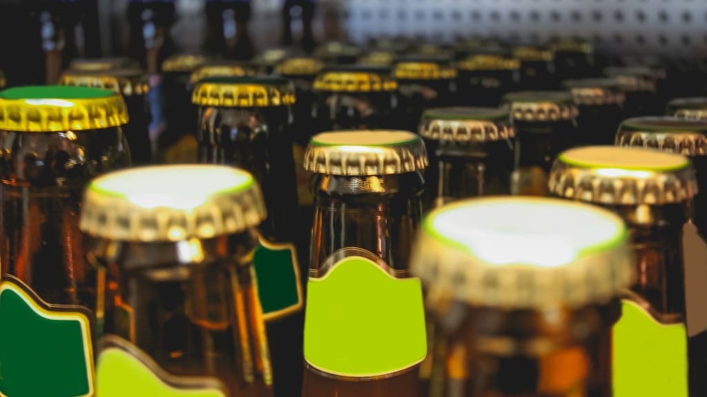 Several brown glass beer bottles with blank green labels and gold caps are arranged closely together on a shelf, ready for sale under an off-premise license.